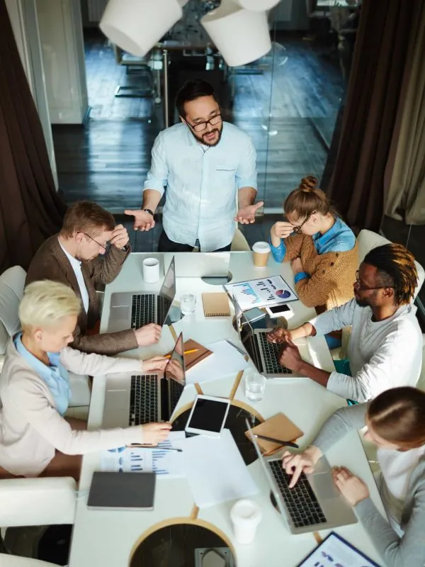 Project team working around a meeting table with laptops while a colleague stands to speak